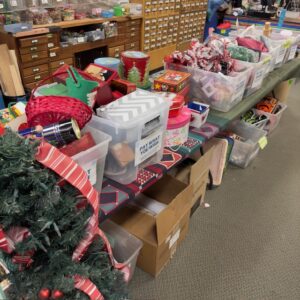 Tables with bins on top and below filled with various holiday decorations, ribbons, gift boxes, etc.